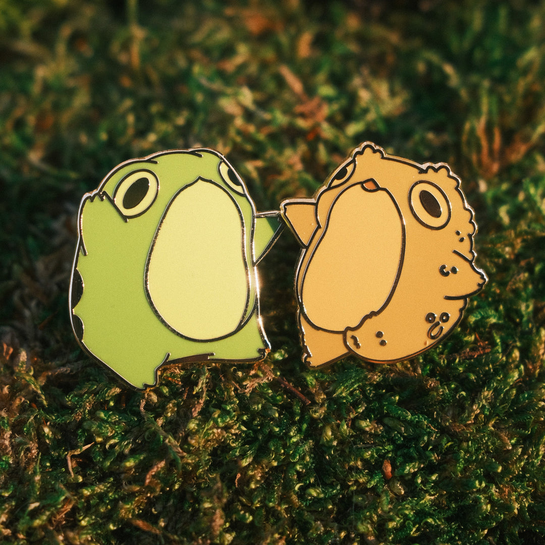 two pins of a frog and toad dancing together on a mossy background