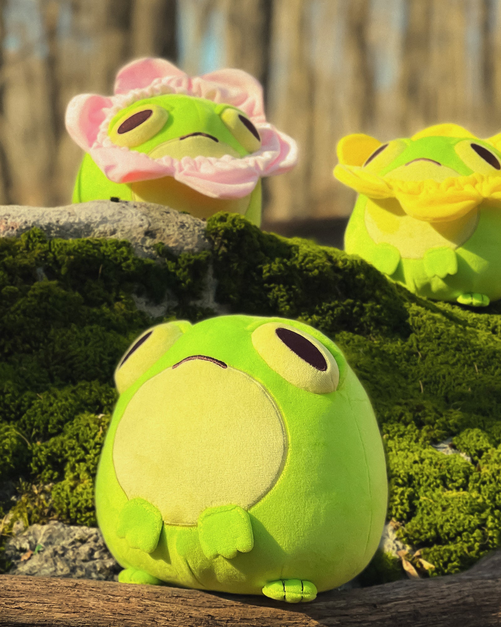 Three green plush toys resembling frogs with a flower necklace in a mossy forest
