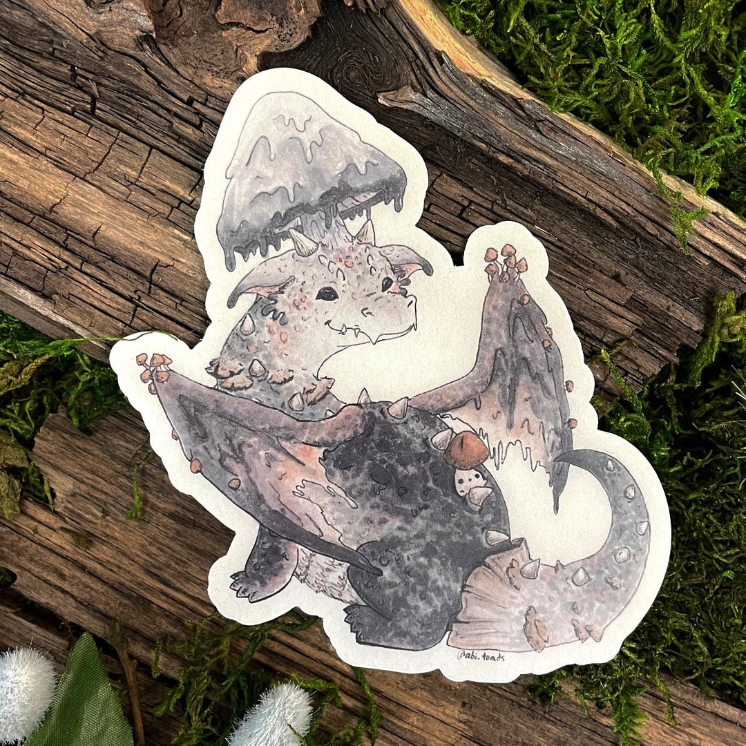 A vinyl sticker depicting a cute, cartoon-style mushroom dragon with a fantasy theme, placed on a wooden surface with moss.