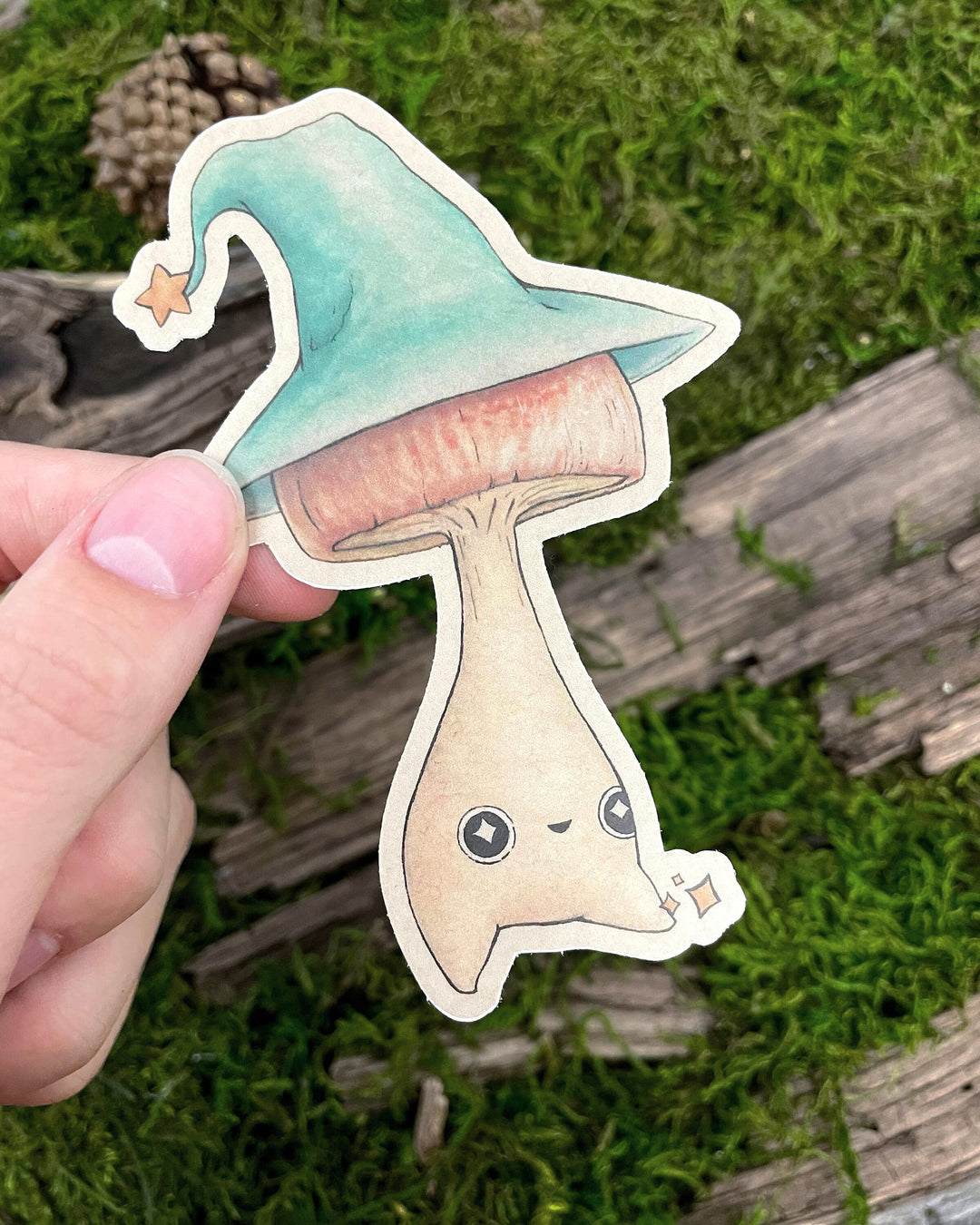 A sticker depicting a cute mushroom wearing a wizard's hat held by a hand