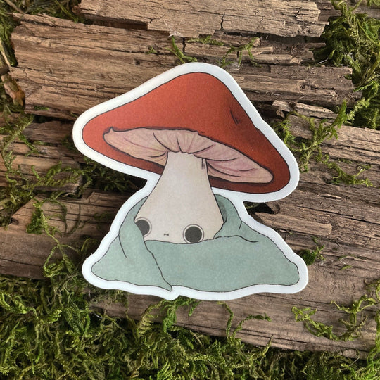 A sticker depicting a cute, cozy mushroom in a blanket position, with a vibrant red cap and a gentle eye design, placed on a wooden surface with green moss.