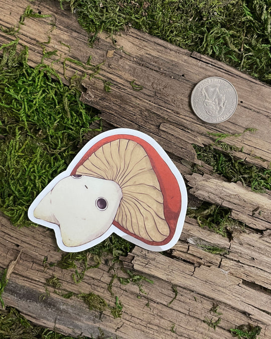 A clear sticker with a illustration of a mushroom character on a wooden surface with green moss next to a quarter for reference