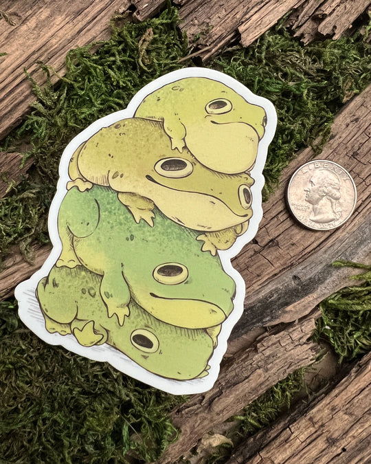 A vinyl sticker depicting a group of green toad stickers stacked on top of each other, placed on a wooden surface with moss next to a quarter for reference