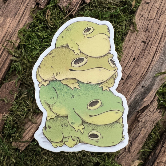 A vinyl sticker depicting a group of green toad stickers stacked on top of each other, placed on a wooden surface with moss.