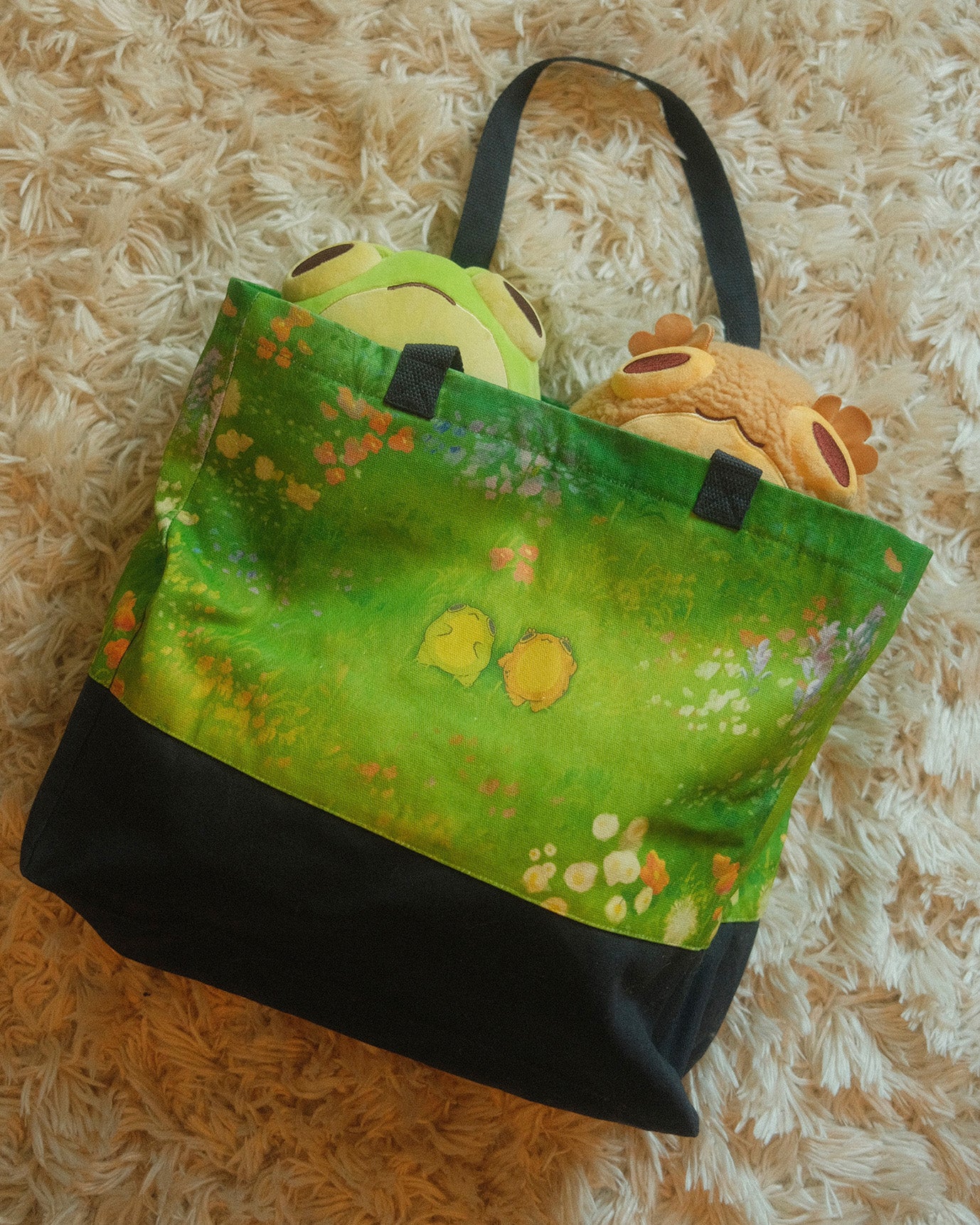 tote bag depicting two frogs laying in a flowery field, with two frog plushies in the bag